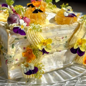 A very close up of a brie square that has been cut in have and layered with a whipped radish butter brie. The brie cake is then frosted with more whipped butter and decorated with mini radishes, edible flowers and honeycomb on a crystal plate.
