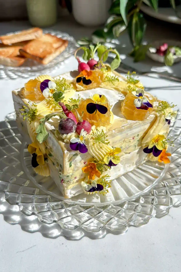 Saint Angel brie square that has been cut in have and layered with a whipped radish butter brie. The brie cake is then frosted with more whipped butter and decorated with mini radishes, edible flowers and honeycomb on a crystal plate.