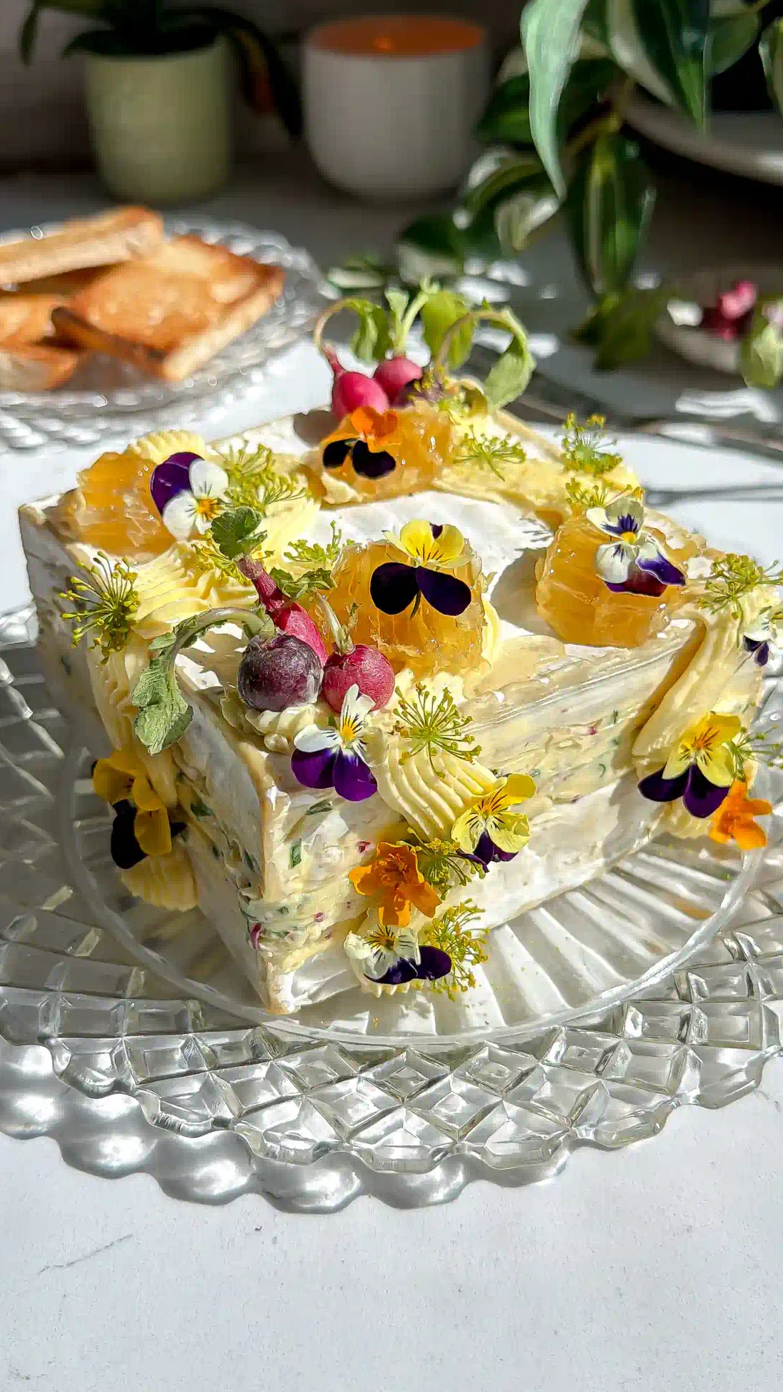 Saint Angel brie square that has been cut in have and layered with a whipped radish butter brie. The brie cake is then frosted with more whipped butter and decorated with mini radishes, edible flowers and honeycomb on a crystal plate.