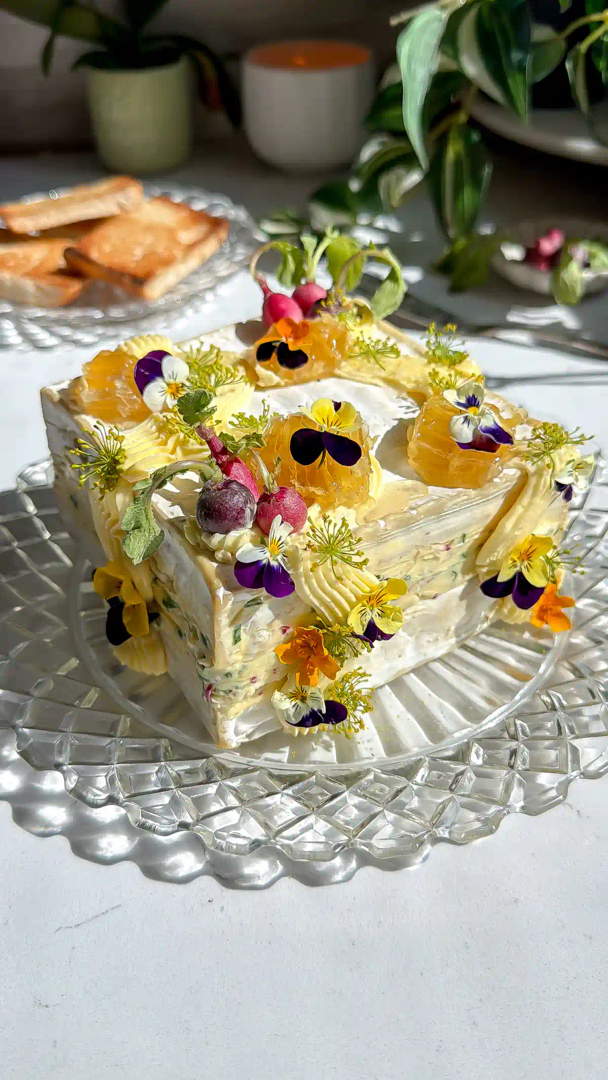 Saint Angel brie square that has been cut in have and layered with a whipped radish butter brie. The brie cake is then frosted with more whipped butter and decorated with mini radishes, edible flowers and honeycomb on a crystal plate.