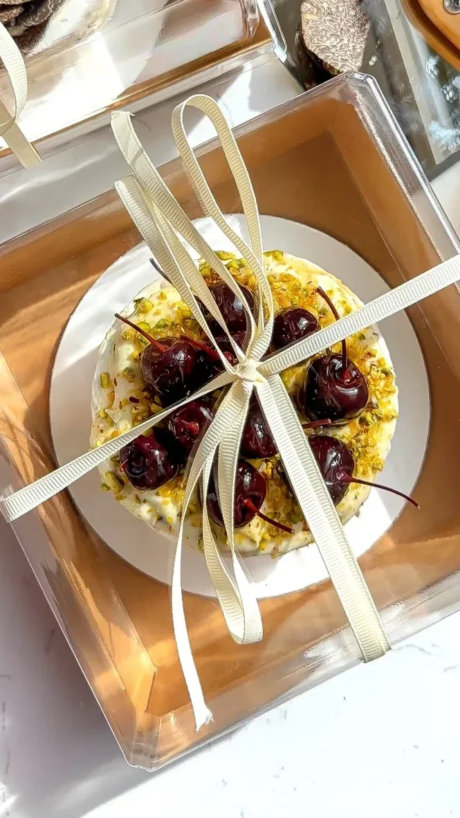 Overhead view of brie cake with whipped cherry brie, pistachios, and whole cherries tied with ribbon in a gift box.