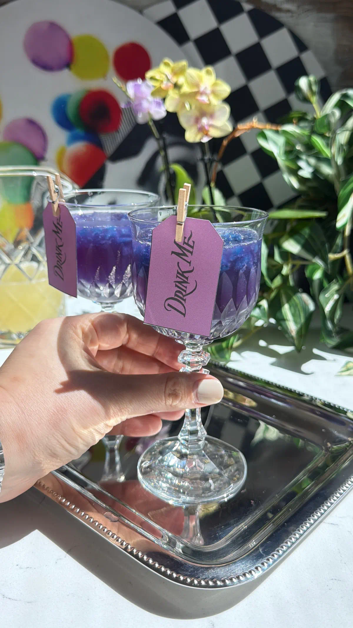 A hand holding a crystal glass filled with a purple color-changing galaxy cocktail over shaved blue butterfly pea granita, featuring a Drink Me tag.