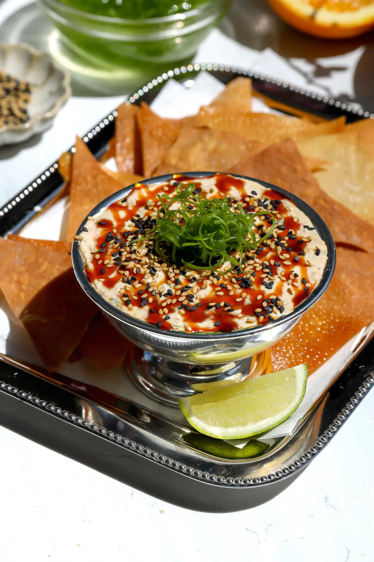 A close-up of cold deconstructed crab rangoon dip in a silver pedestal bowl, topped with a glossy chili honey reduction, black and white sesame seeds, and curled scallions, served on a silver tray with crispy baked wonton chips.