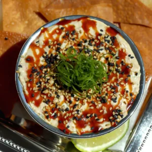 An overhead top-down view of a silver bowl filled with cold crab rangoon dip, highlighting the detailed garnish of drizzled chili honey, sesame seeds, and a center pile of curled green scallions.