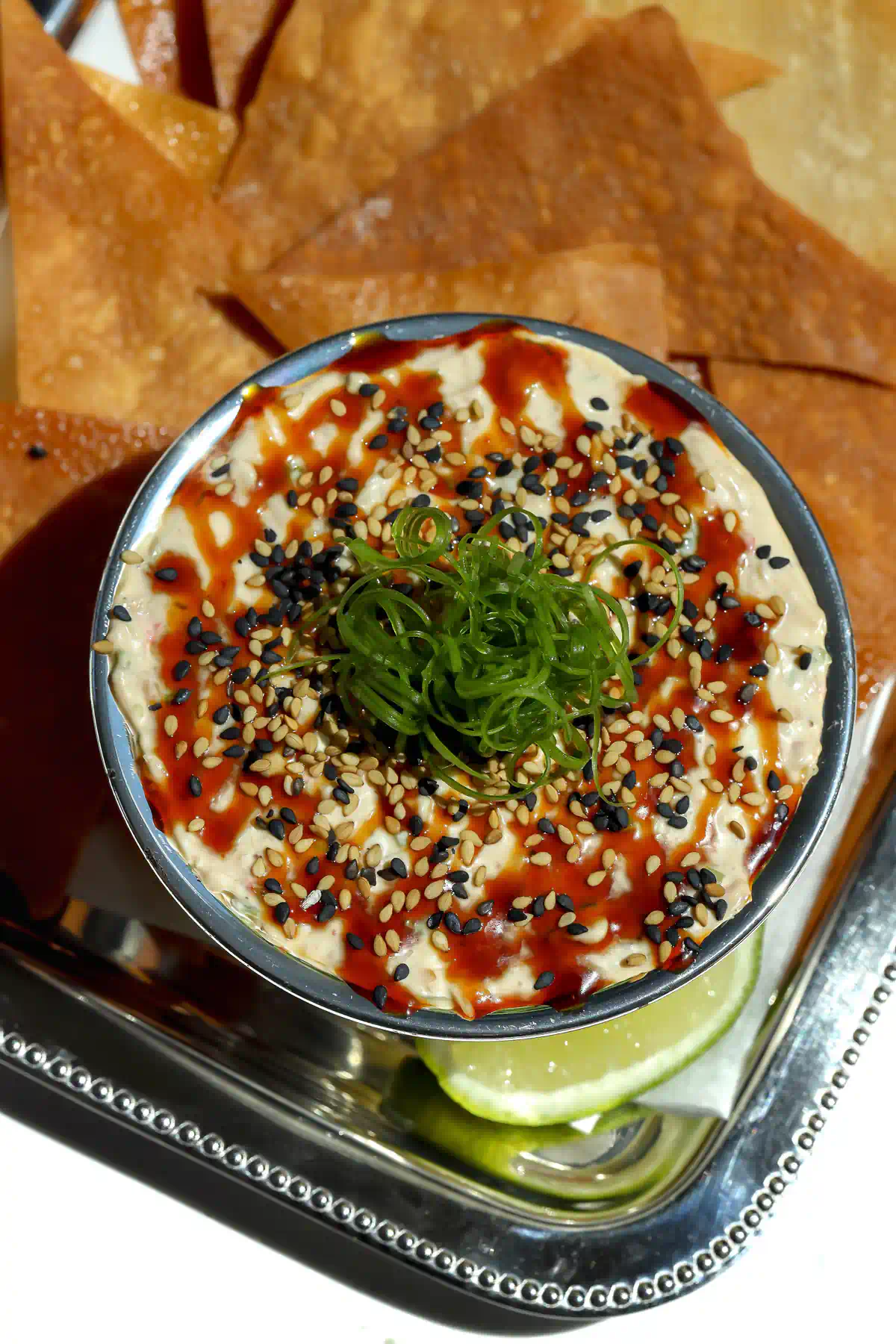 An overhead top-down view of a silver bowl filled with cold crab rangoon dip, highlighting the detailed garnish of drizzled chili honey, sesame seeds, and a center pile of curled green scallions.