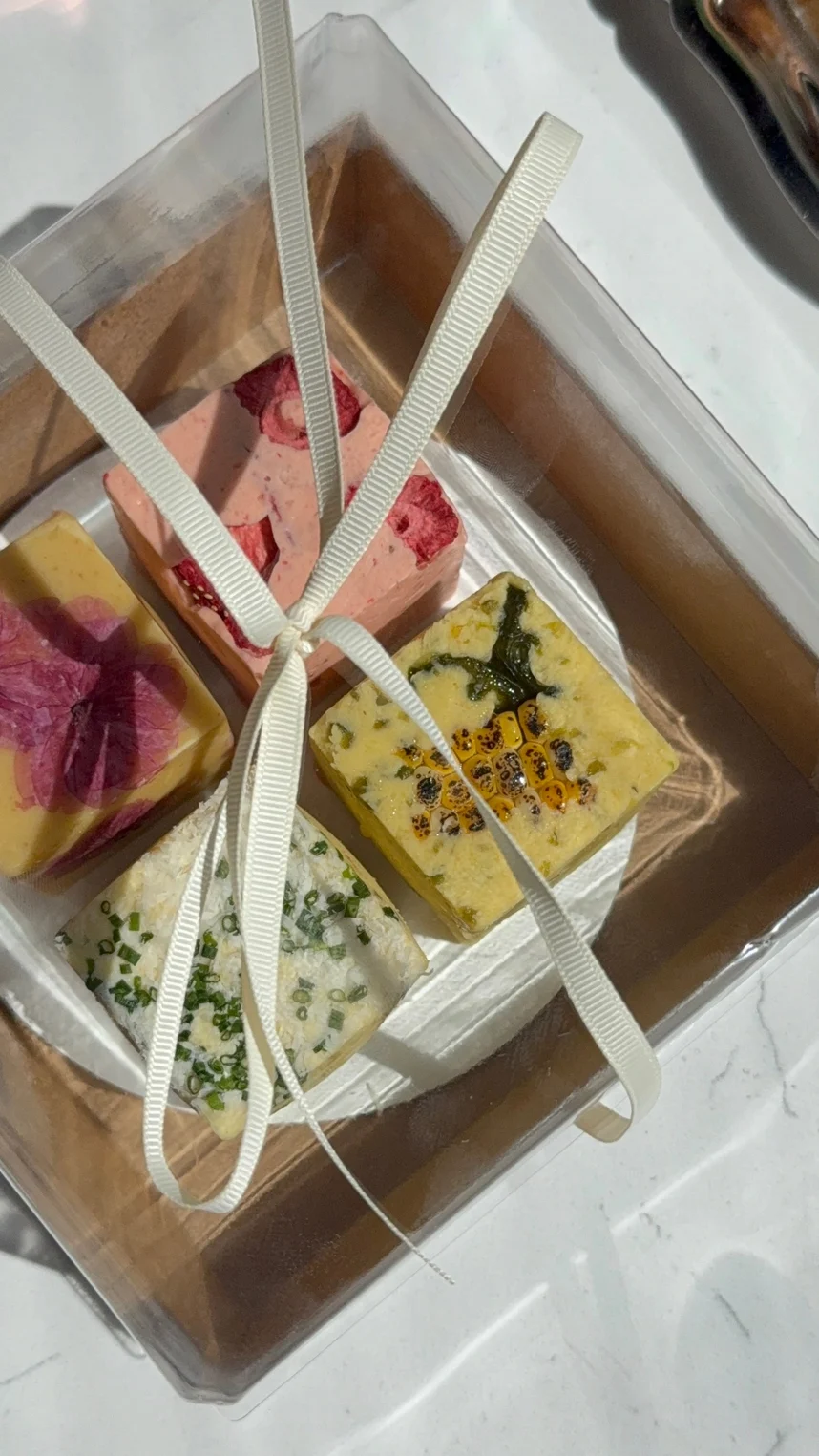 Four unwrapped blocks of summer compound butter arranged in a clear square gift box tied with a white ribbon.
