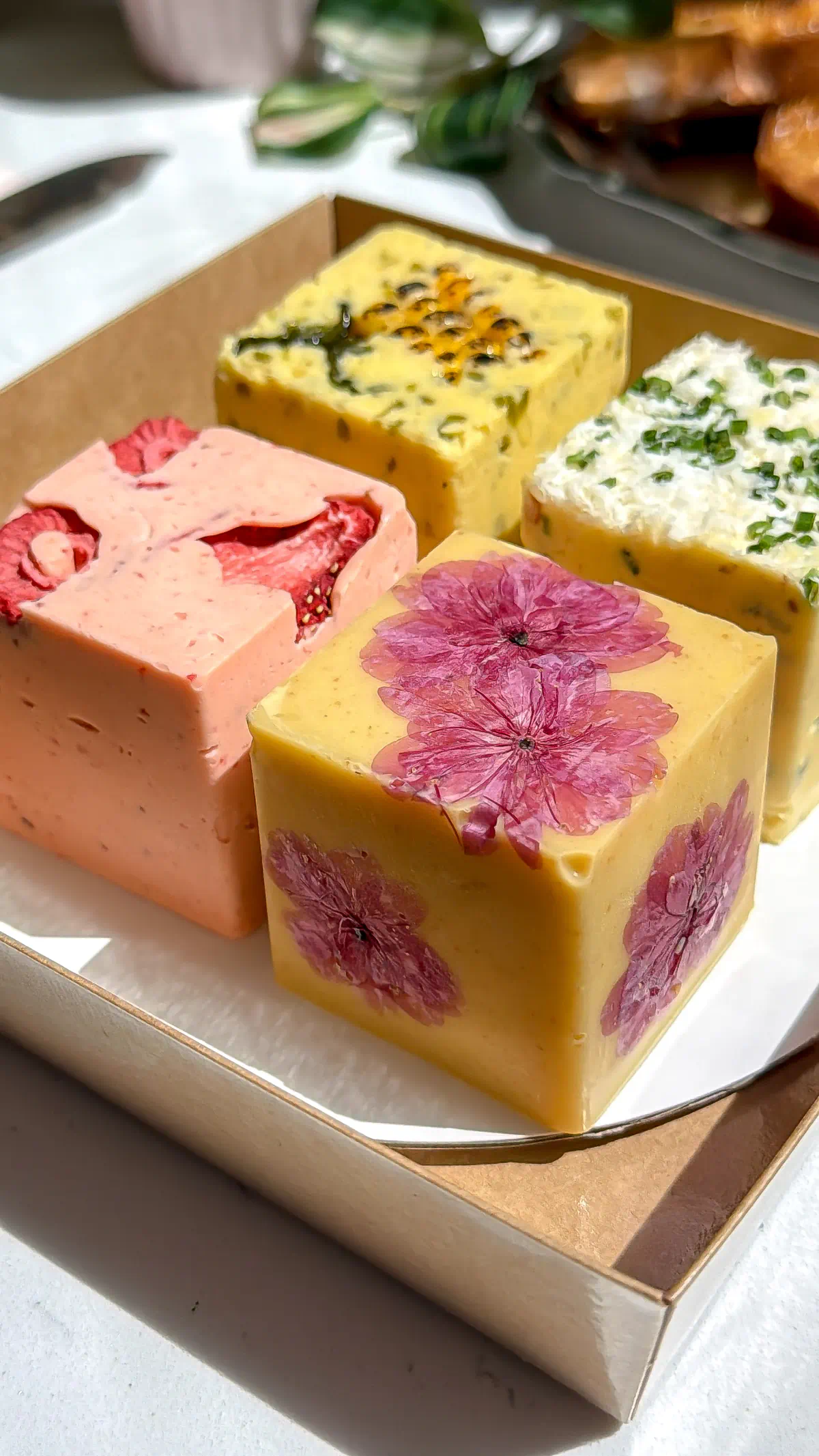 Four square blocks of a summer compound butter flight in a box, featuring strawberry, pressed cherry blossom, jalape&ntilde;o corn, and garlic chive flavors.