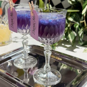 Two crystal glasses filled with purple galaxy cotton candy lemonade cocktails over blue granita, resting on a silver tray with Drink Me tags attached.