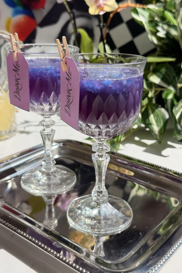 Two crystal glasses filled with purple galaxy cotton candy lemonade cocktails over blue granita, resting on a silver tray with Drink Me tags attached.