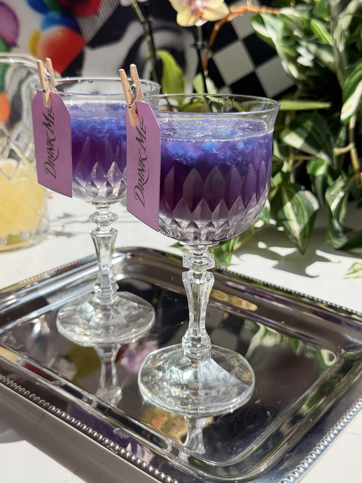Two crystal glasses filled with purple galaxy cotton candy lemonade cocktails over blue granita, resting on a silver tray with Drink Me tags attached.