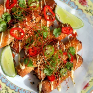 Top-down view of golden fried crab Rangoon taquitos on a parchment-lined plate, topped with lemon mayo, sweet chili honey, sliced jalapenos, red chiles, fresh cilantro, and sesame seeds.