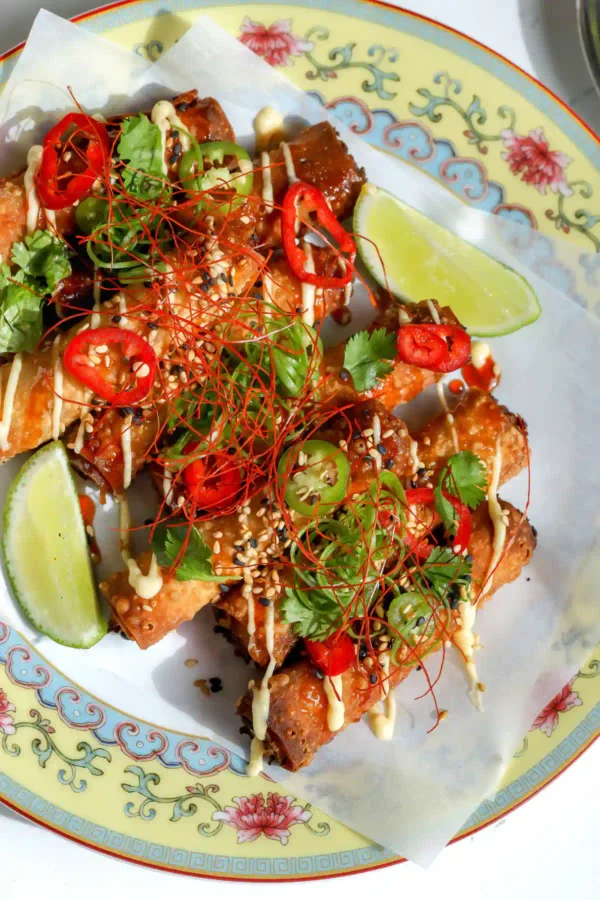 Top-down view of golden fried crab Rangoon taquitos on a parchment-lined plate, topped with lemon mayo, sweet chili honey, sliced jalapenos, red chiles, fresh cilantro, and sesame seeds.