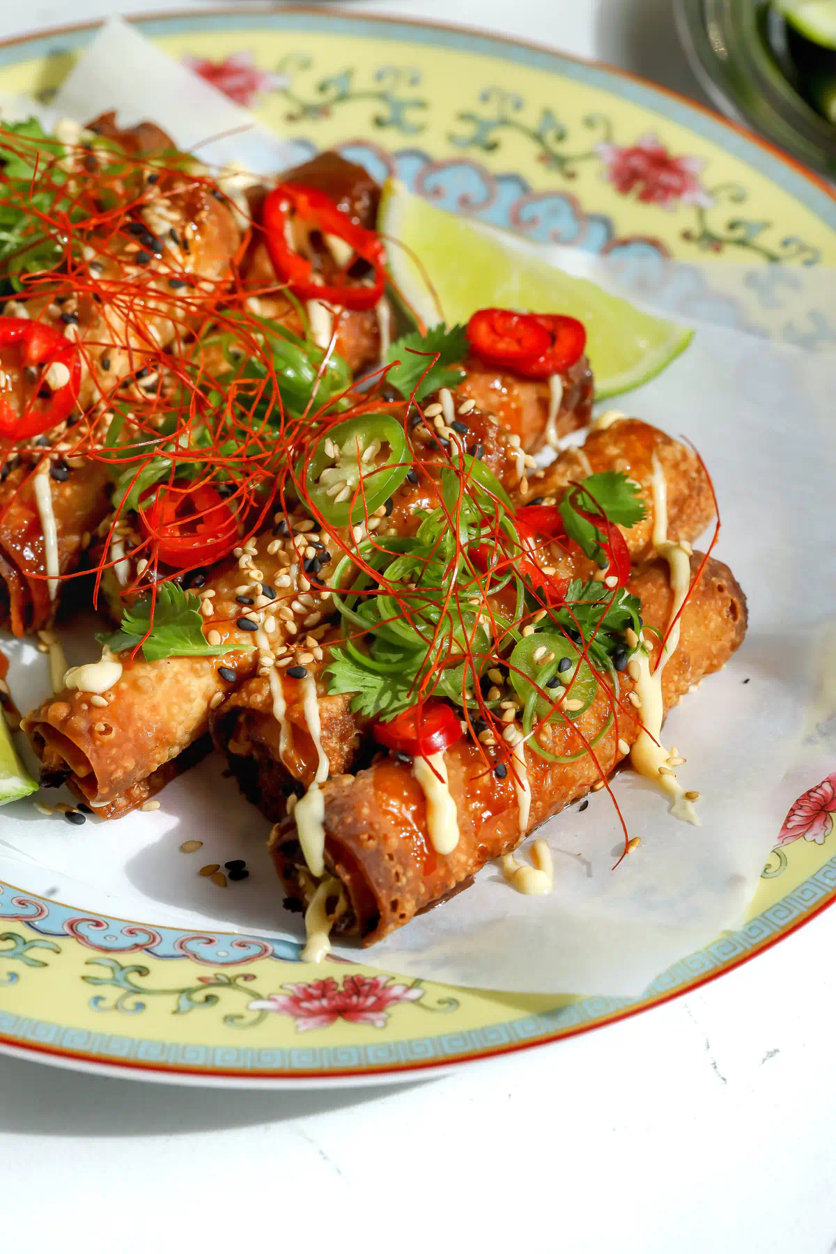 Close-up side view of crispy crab Rangoon taquitos piled on a decorative plate, drizzled with sweet chili honey and lemon mayo, and garnished with sesame seeds, cilantro, sliced chiles, and red chili threads.