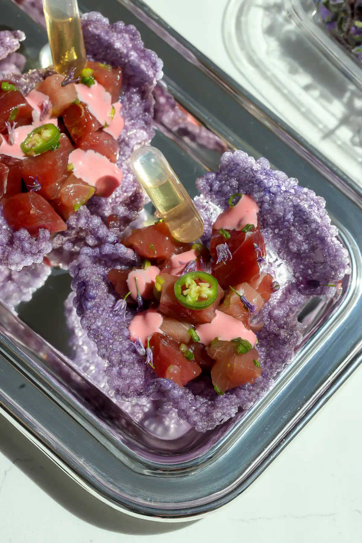 Overhead view of purple tapioca crisps filled with cured bluefin tuna, pink mayo, and a shiro dashi ponzu syringe on a silver platter.