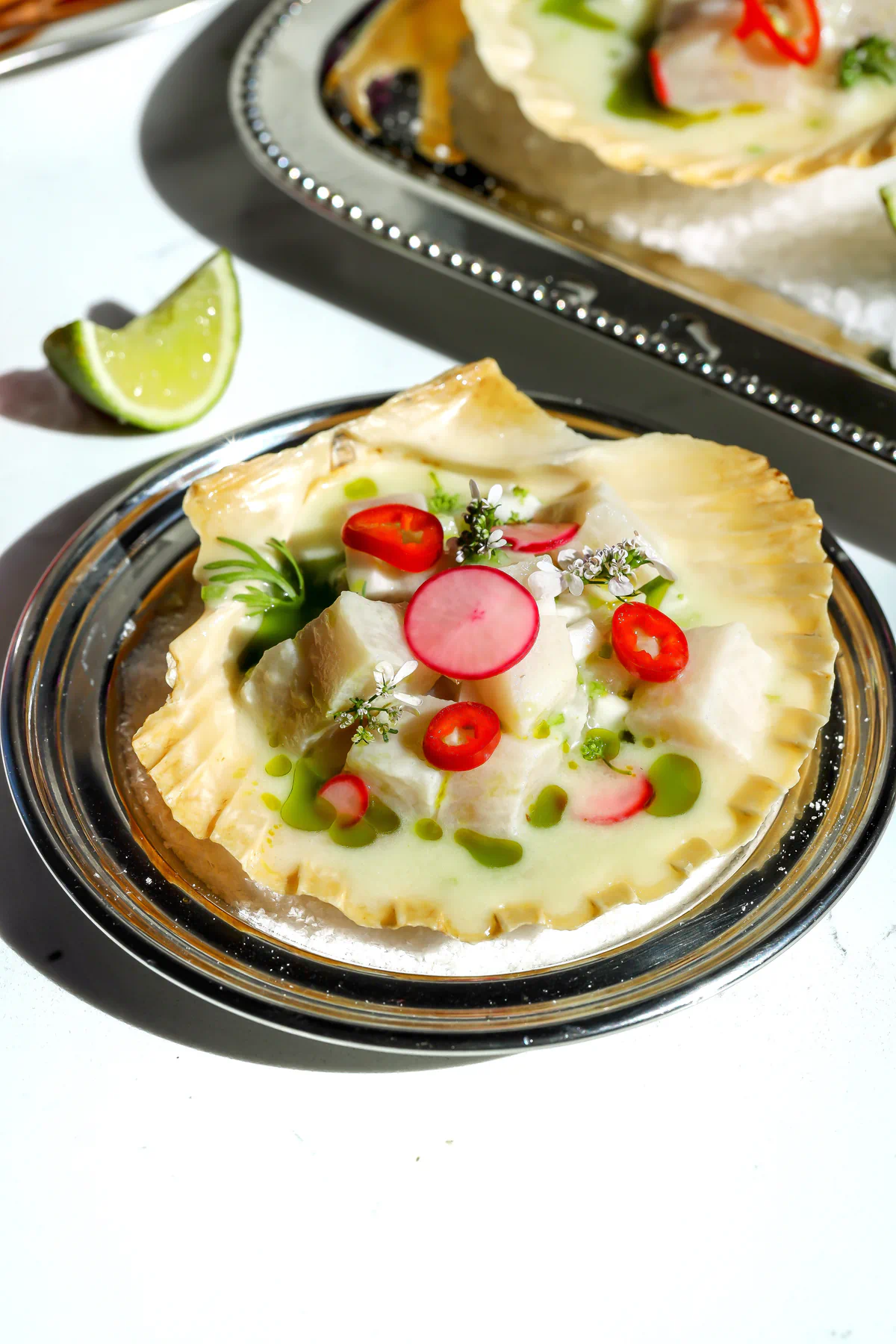 Yellowtail ceviche served in a scallop shell with coconut leche de tigre, cilantro oil, pickled radish, and sliced red chili