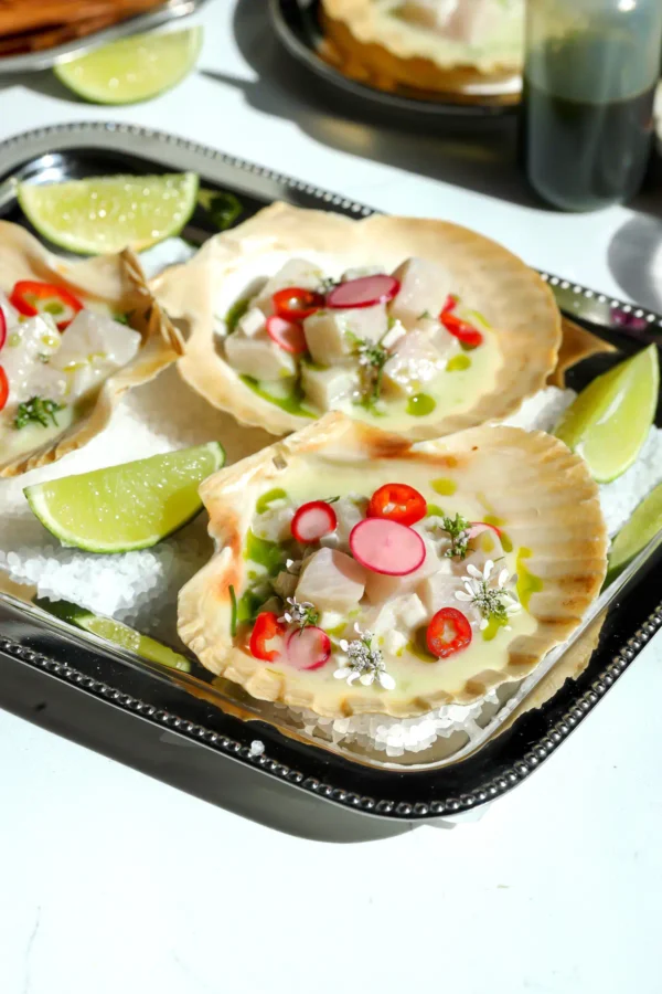 Hamachi yellowtail ceviche in scallop shells arranged on a silver serving tray with lime wedges and cilantro oil.