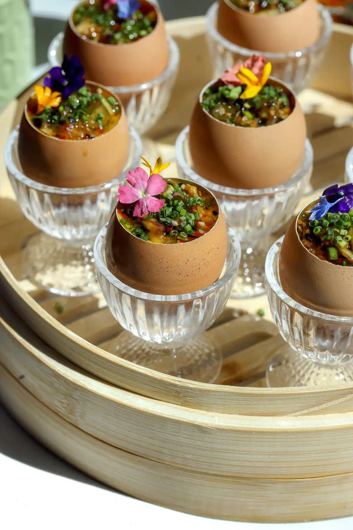Six chawanmushi steamed egg custards served in brown egg shells topped with shallot chili crisp and colorful edible flowers, sitting in crystal egg cups on a bamboo steamer tray