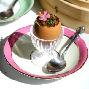 Single serving of chawanmushi in a brown egg shell topped with shallot chili crisp and a pink edible flower, in a crystal egg cup on a pink and gold rimmed plate with a silver spoon