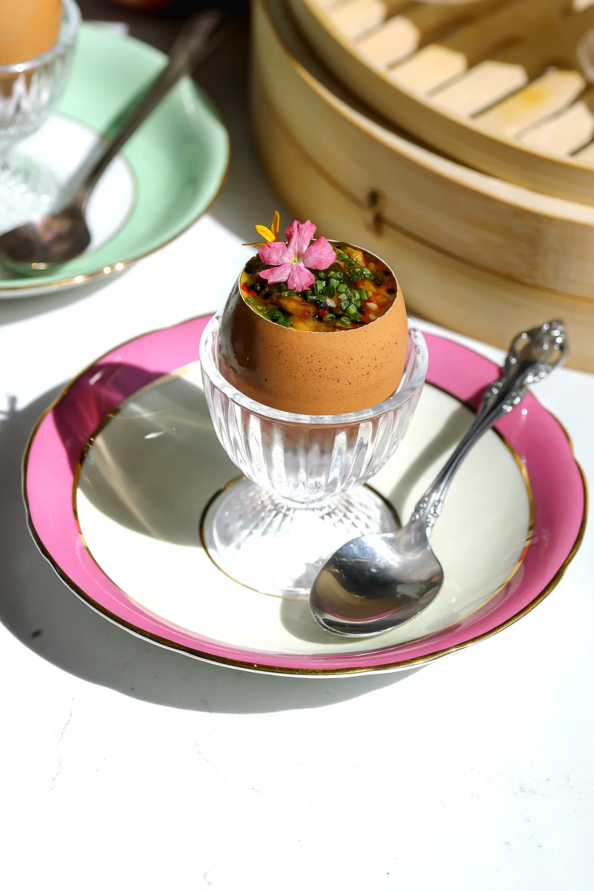 Single serving of chawanmushi in a brown egg shell topped with shallot chili crisp and a pink edible flower, in a crystal egg cup on a pink and gold rimmed plate with a silver spoon