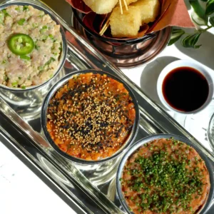 Crispy rice tartare flight with yellowtail, spicy tuna, and salmon tartares in silver bowls next to a basket of crispy rice bites on toothpicks and a small bowl of eel sauce.