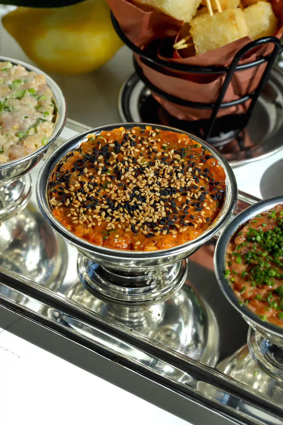 Salsa macha salmon tartare topped with black and white sesame seeds in a silver footed bowl, with the basket of crispy rice bites behind it.
