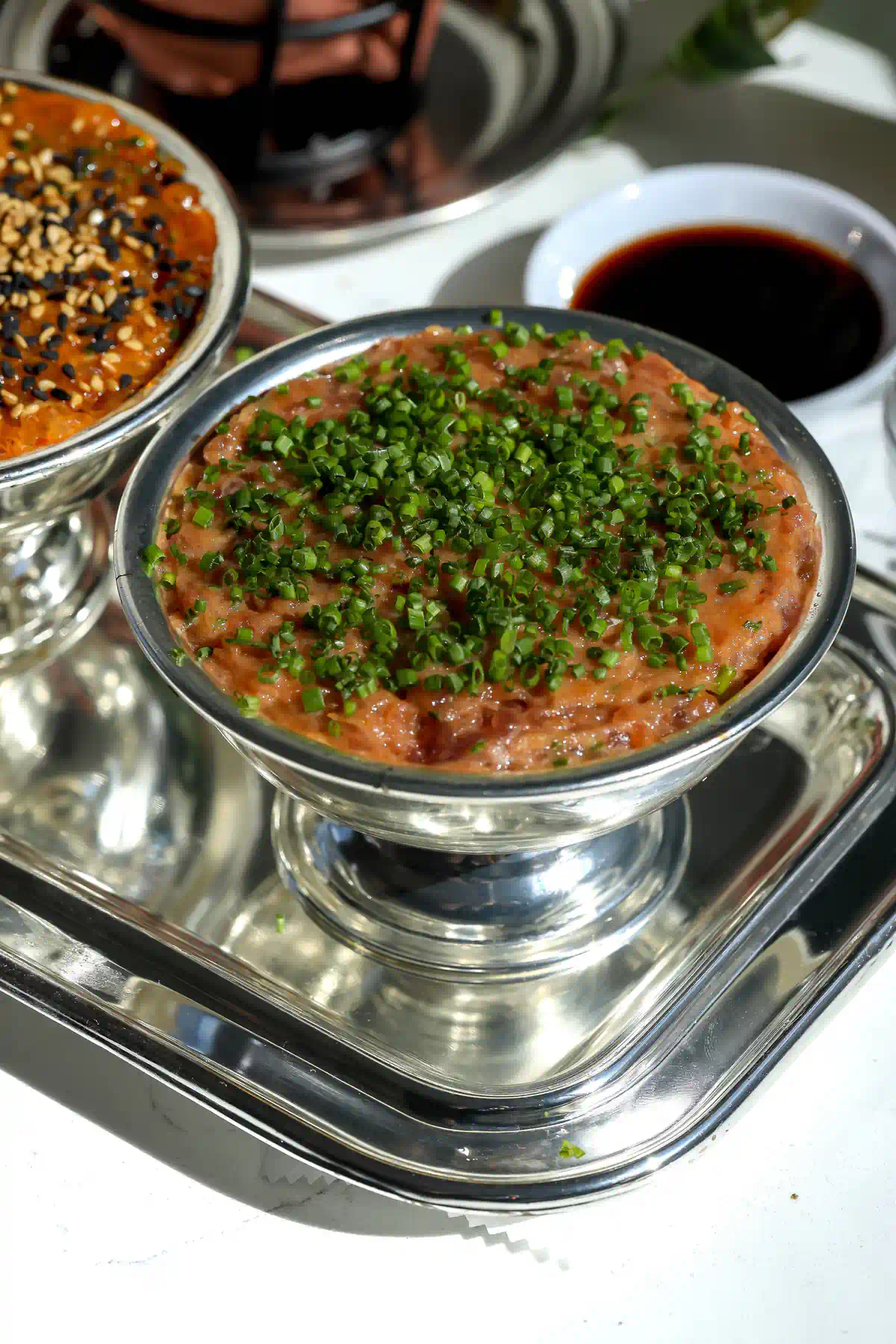 Spicy tuna tartare topped with chopped chives in a silver footed bowl, with eel sauce and the salmon tartare in the background.