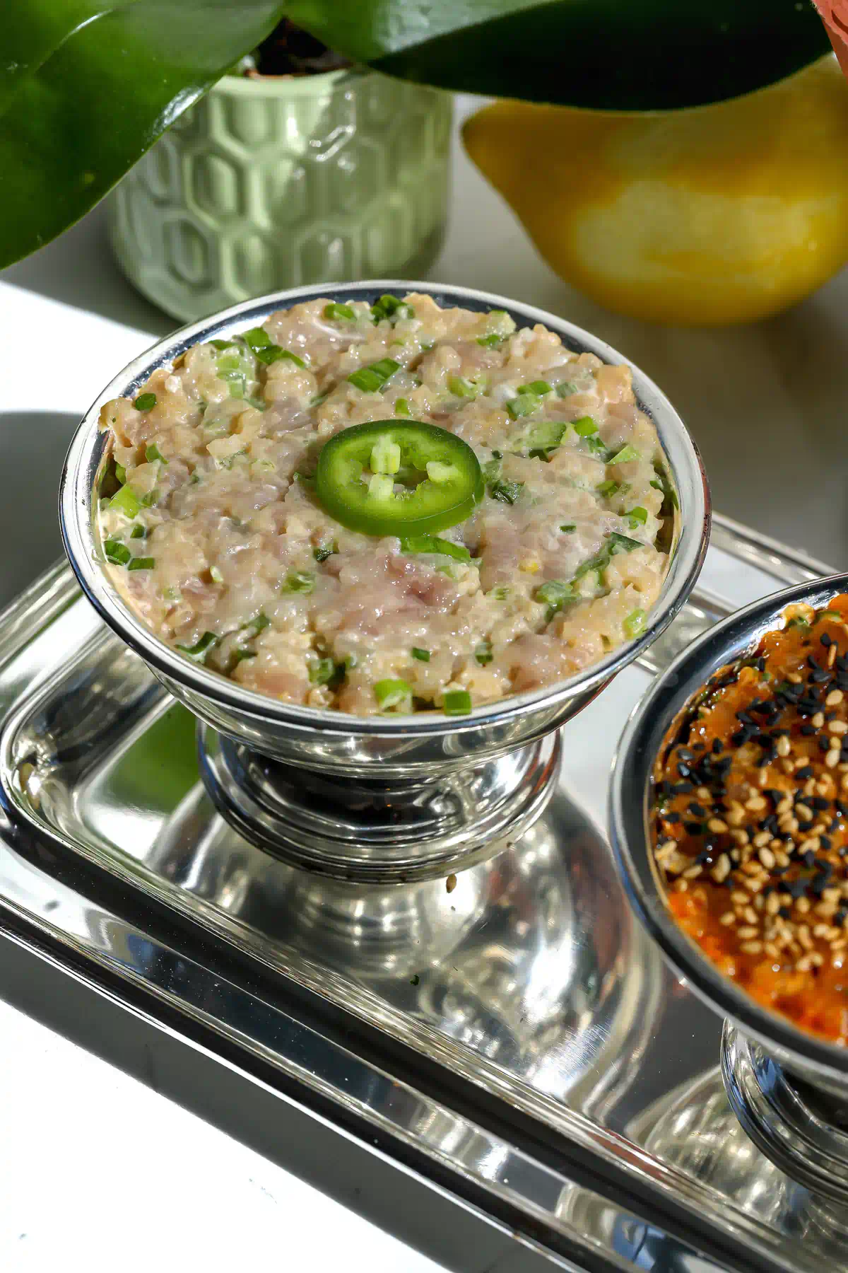 Yellowtail tartare with scallions and a jalapeno slice in a silver footed bowl, with a lemon and the spicy tuna tartare in the background.