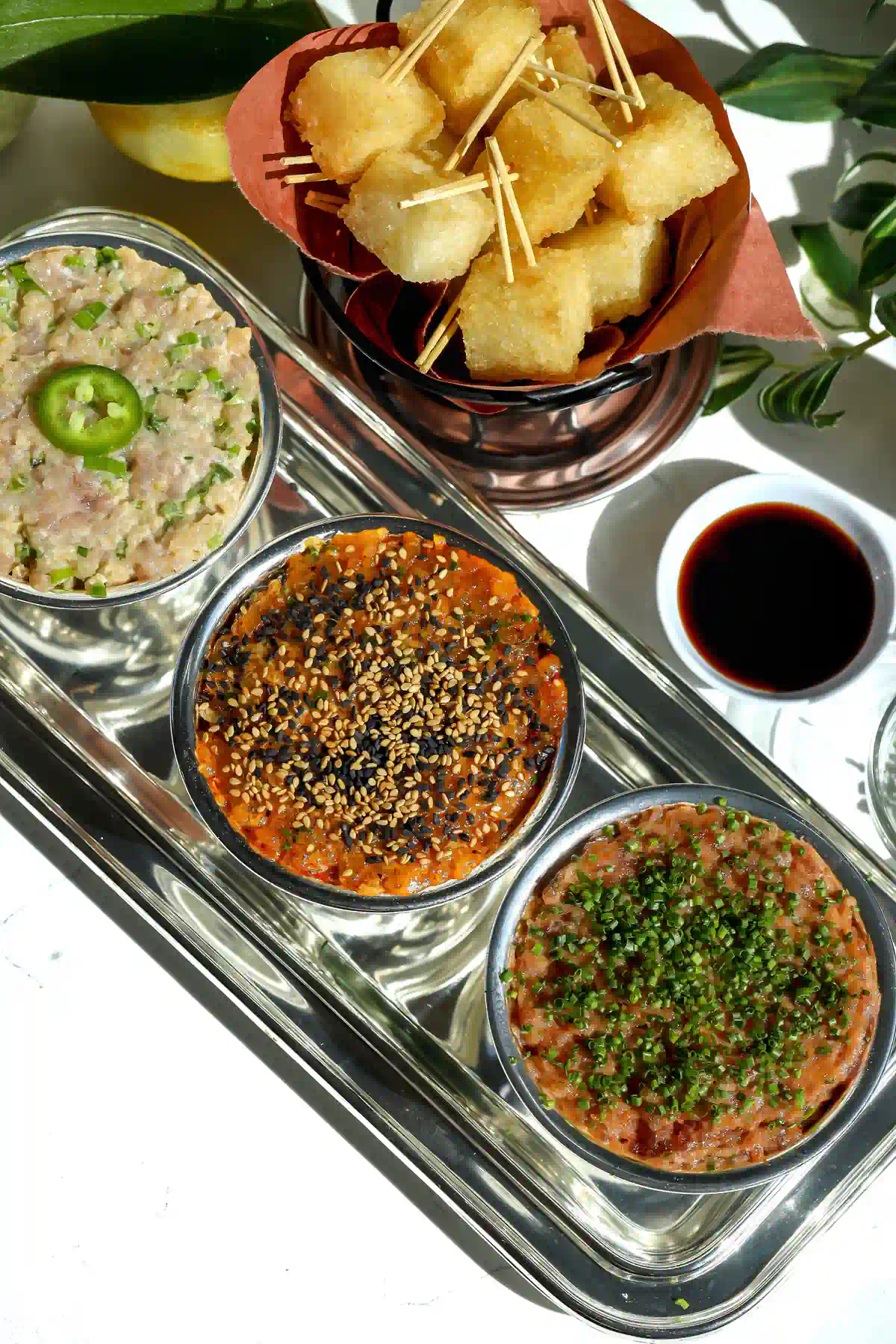 Crispy rice tartare flight with yellowtail, spicy tuna, and salmon tartares in silver bowls next to a basket of crispy rice bites on toothpicks and a small bowl of eel sauce.