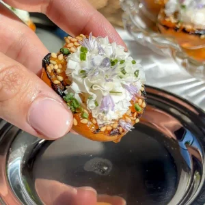 Close-up of a grilled apricot half topped with shaved cheese, toasted sesame seeds, chopped chives, and a purple chive blossom