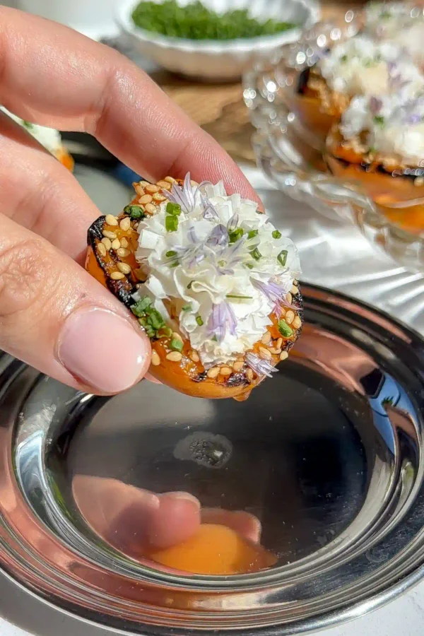 Close-up of a grilled apricot half topped with shaved cheese, toasted sesame seeds, chopped chives, and a purple chive blossom