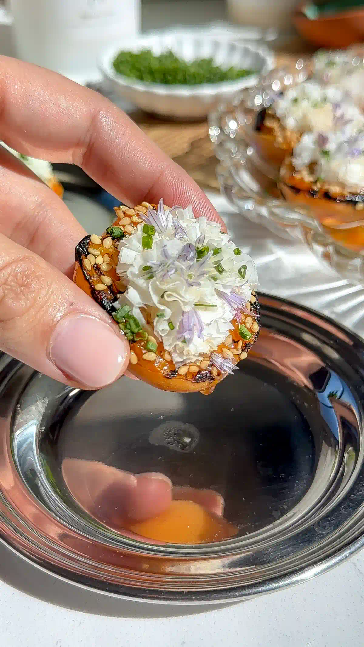 Close-up of a grilled apricot half topped with shaved cheese, toasted sesame seeds, chopped chives, and a purple chive blossom