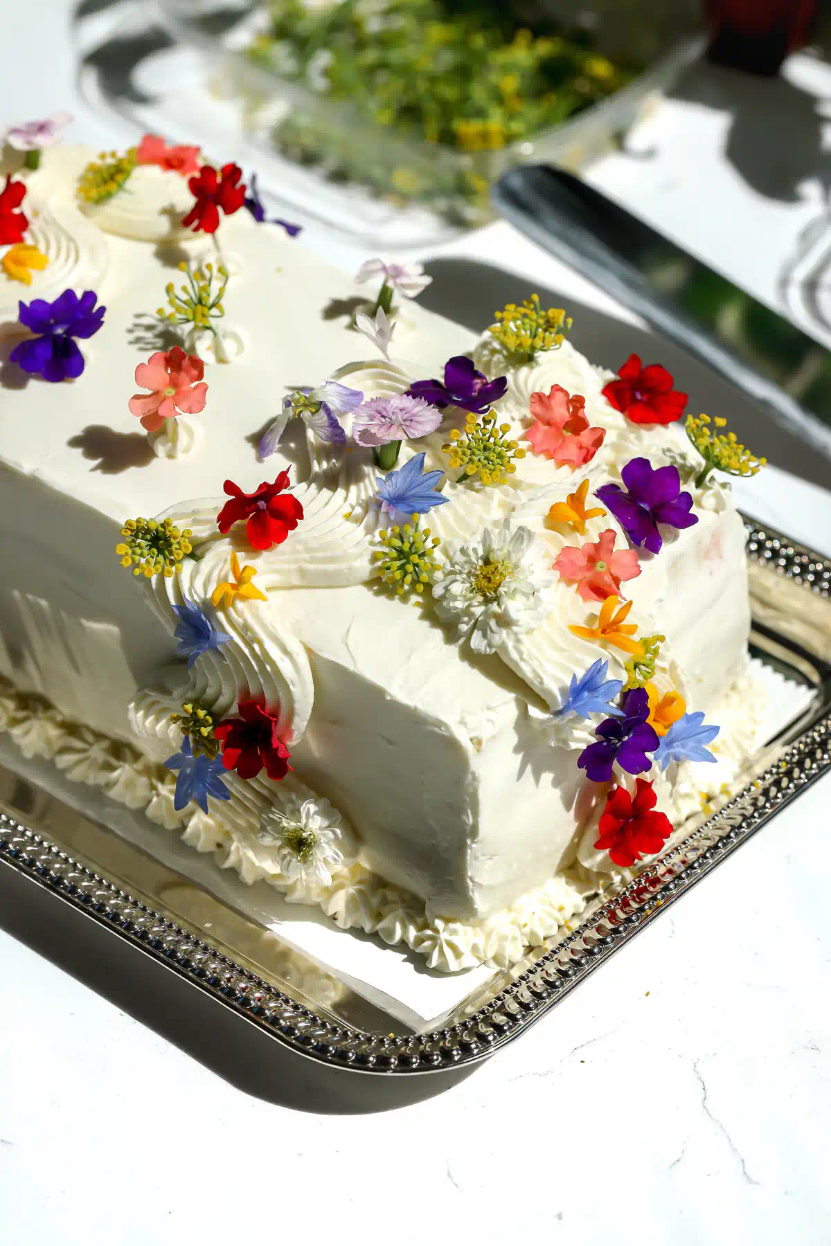 Mexican sandwich cake with white cream cheese frosting topped with red, purple, blue, and orange edible flowers on a silver tray.