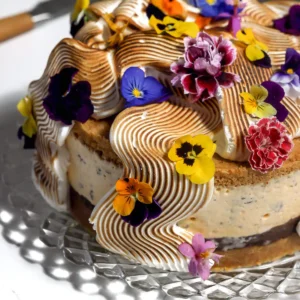 S'mores ice cream cake on a glass cake stand topped with torched Italian meringue in waves and edible flowers, with the chocolate ganache layer and graham crust visible.