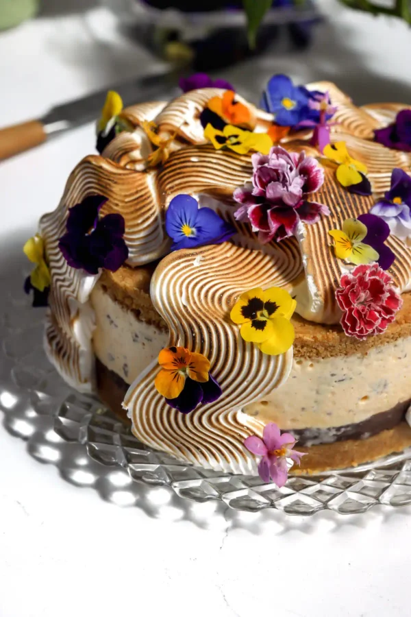 S'mores ice cream cake on a glass cake stand topped with torched Italian meringue in waves and edible flowers, with the chocolate ganache layer and graham crust visible.