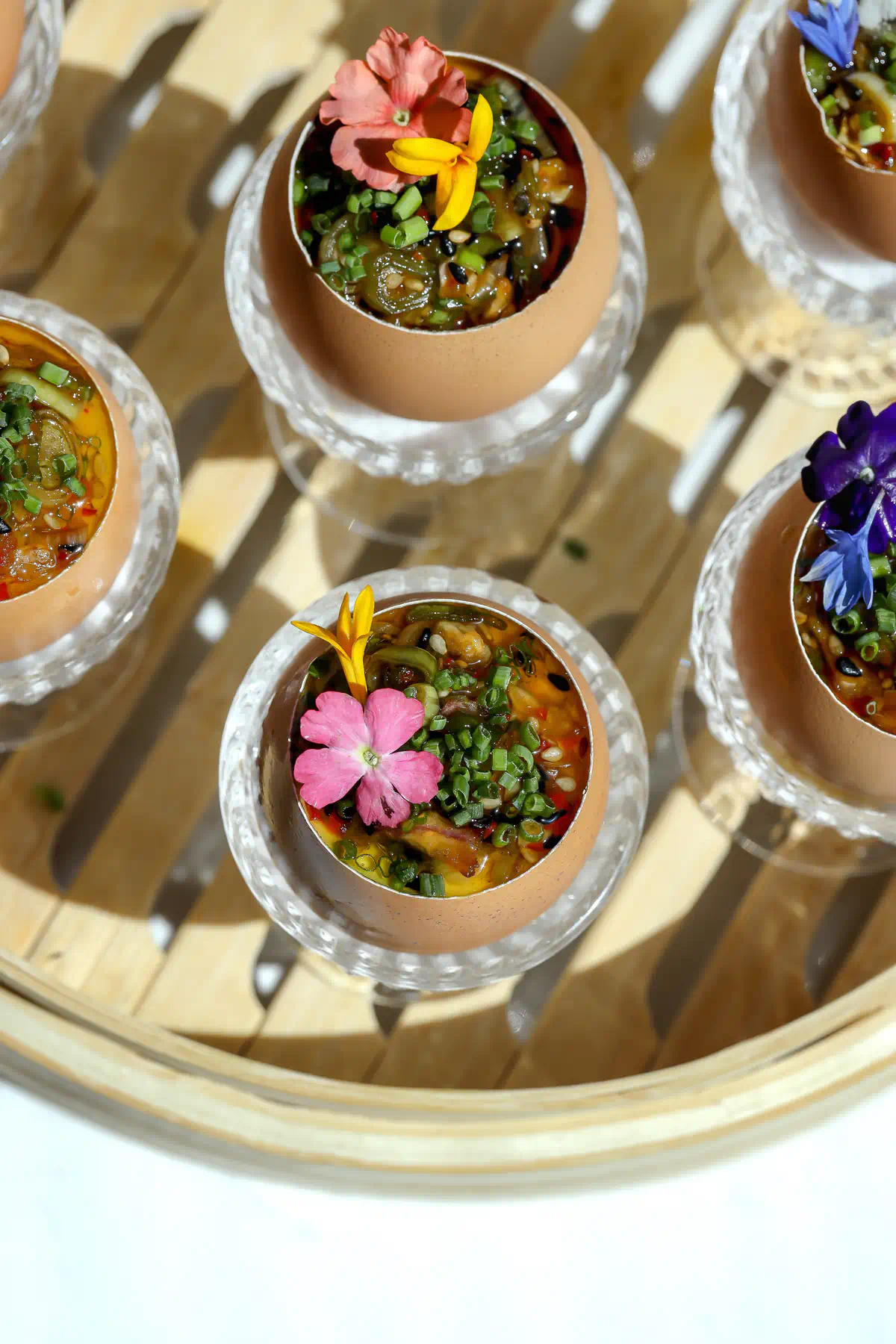 Overhead view of steamed egg custard served in brown egg shells topped with shallot chili crisp and assorted edible flowers in crystal egg cups on a wooden tray.