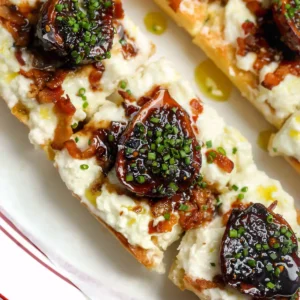 Overhead view of a balsamic fig and stracciatella tartine on toasted baguette, with syrupy braised figs, bacon bits, and chives on a red-rimmed white plate.
