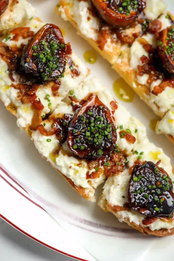 Overhead view of a balsamic fig and stracciatella tartine on toasted baguette, with syrupy braised figs, bacon bits, and chives on a red-rimmed white plate.