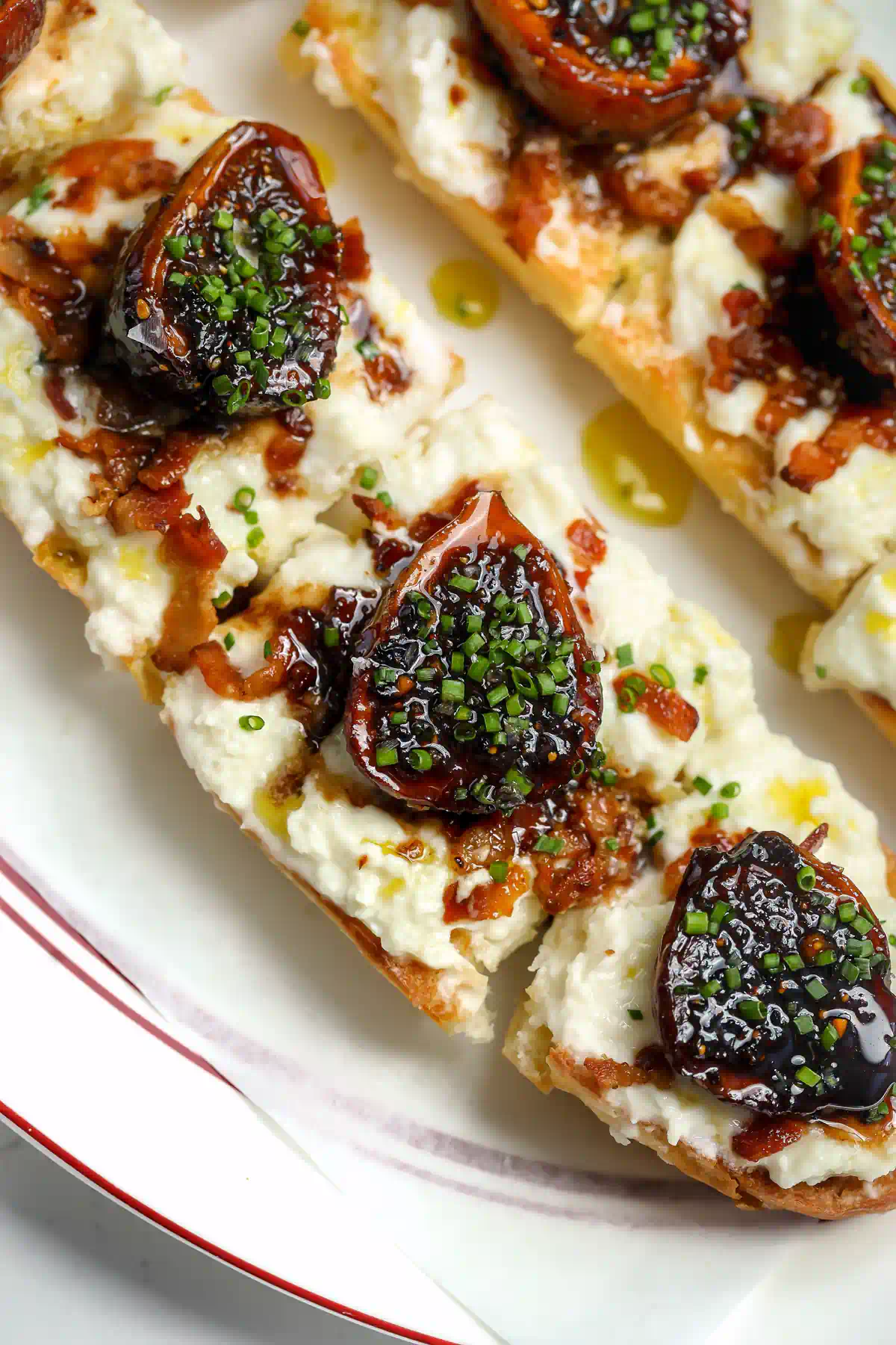 Overhead view of a balsamic fig and stracciatella tartine on toasted baguette, with syrupy braised figs, bacon bits, and chives on a red-rimmed white plate