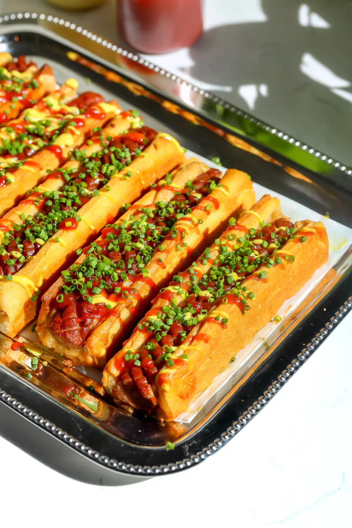 Crescent roll hot dogs on a silver tray topped with spicy ketchup, mustard mayo, and chopped chives.