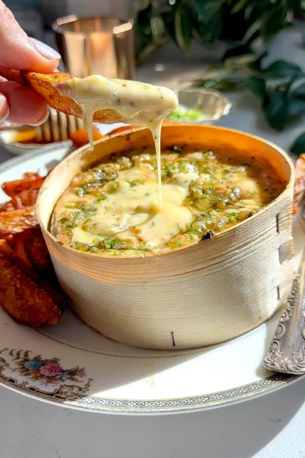 A crispy roasted potato lifting a string of melted cheese from a box of baked Mont d'Or.