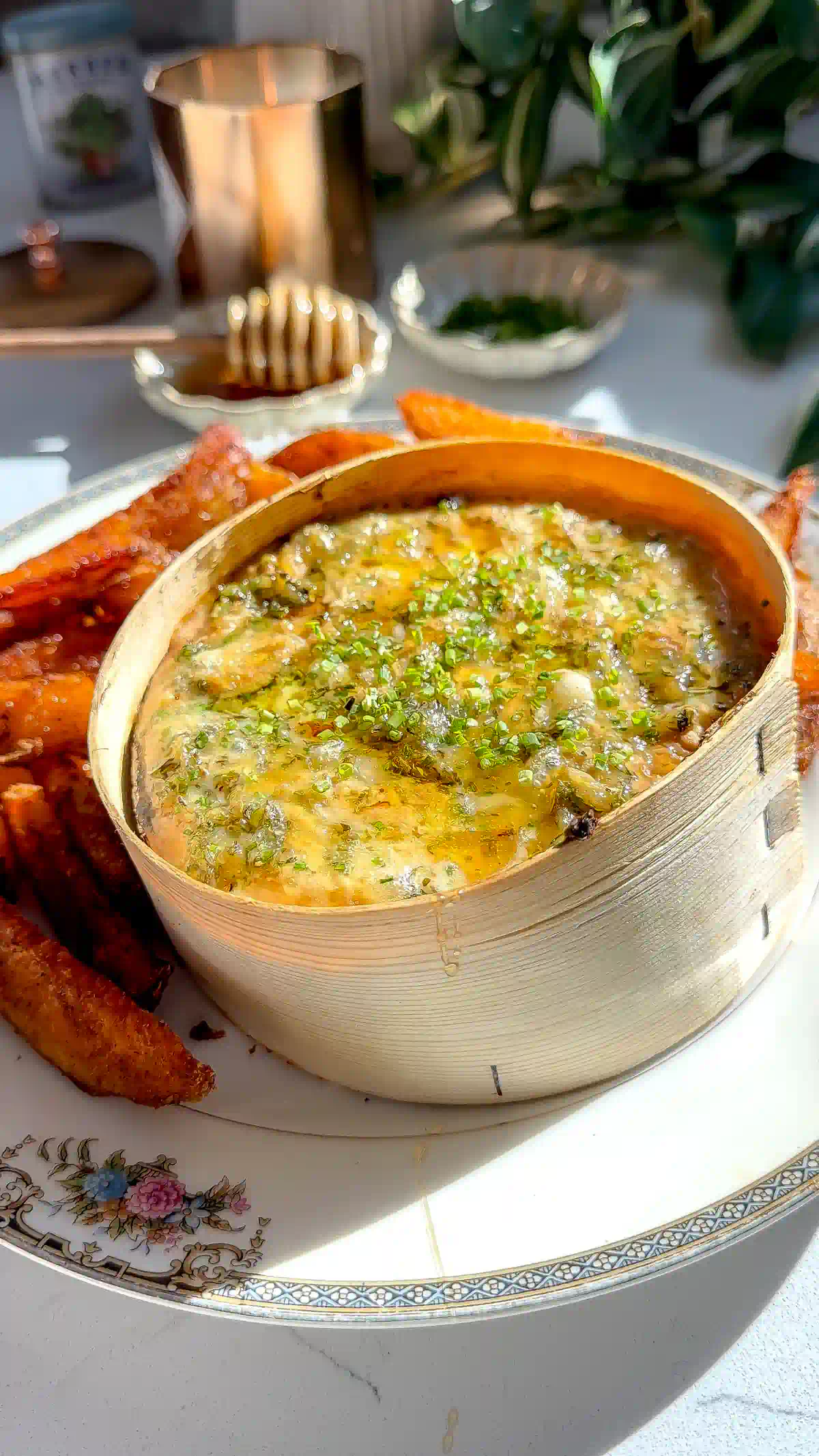 A wheel of baked Mont d'Or in its spruce box topped with melted cheese, roasted garlic herb oil, and chives, surrounded by crispy duck fat roasted potatoes on a floral plate.