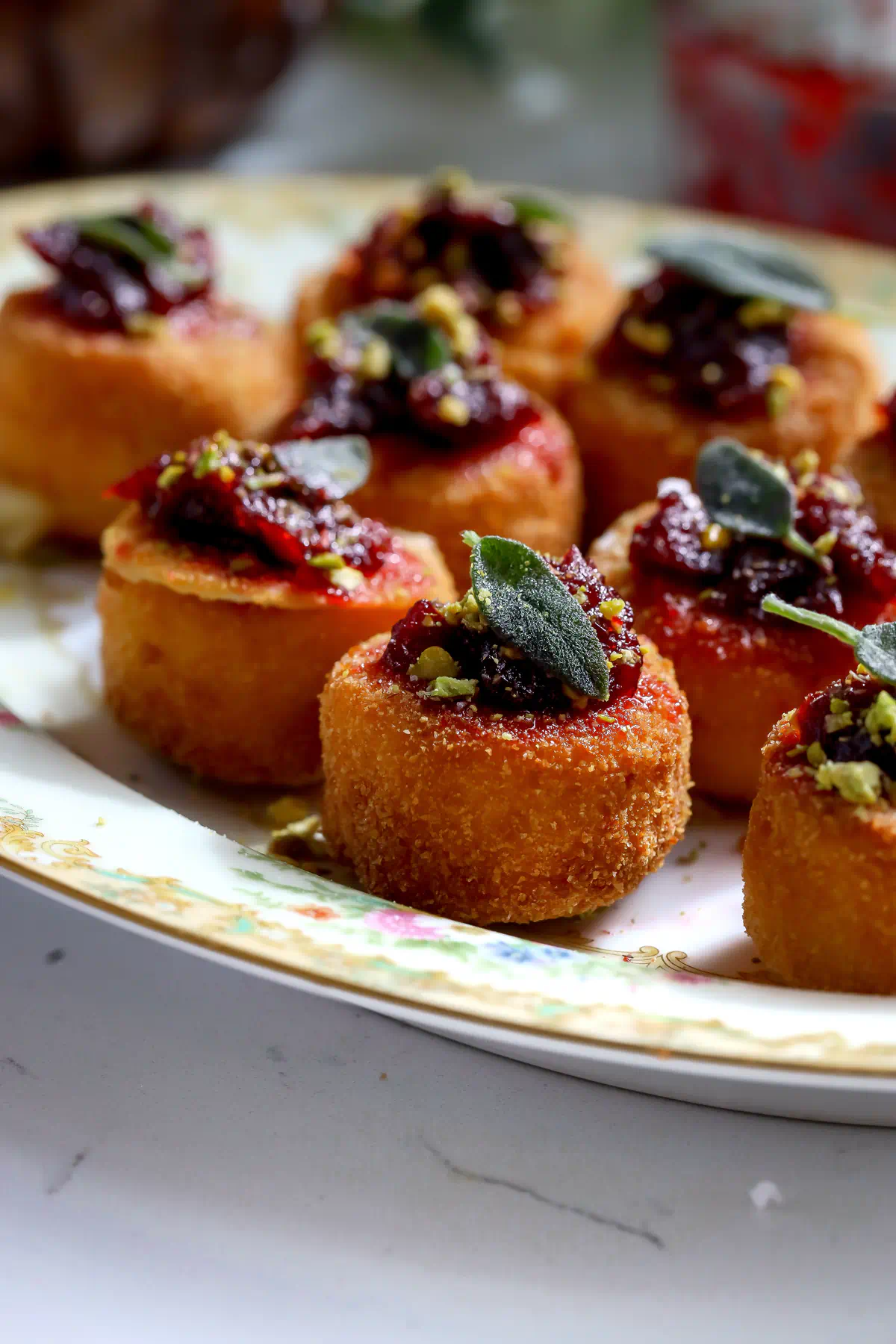 Crispy fried brie bites with cranberry chipotle sauce and fried sage on a gold-rimmed plate.