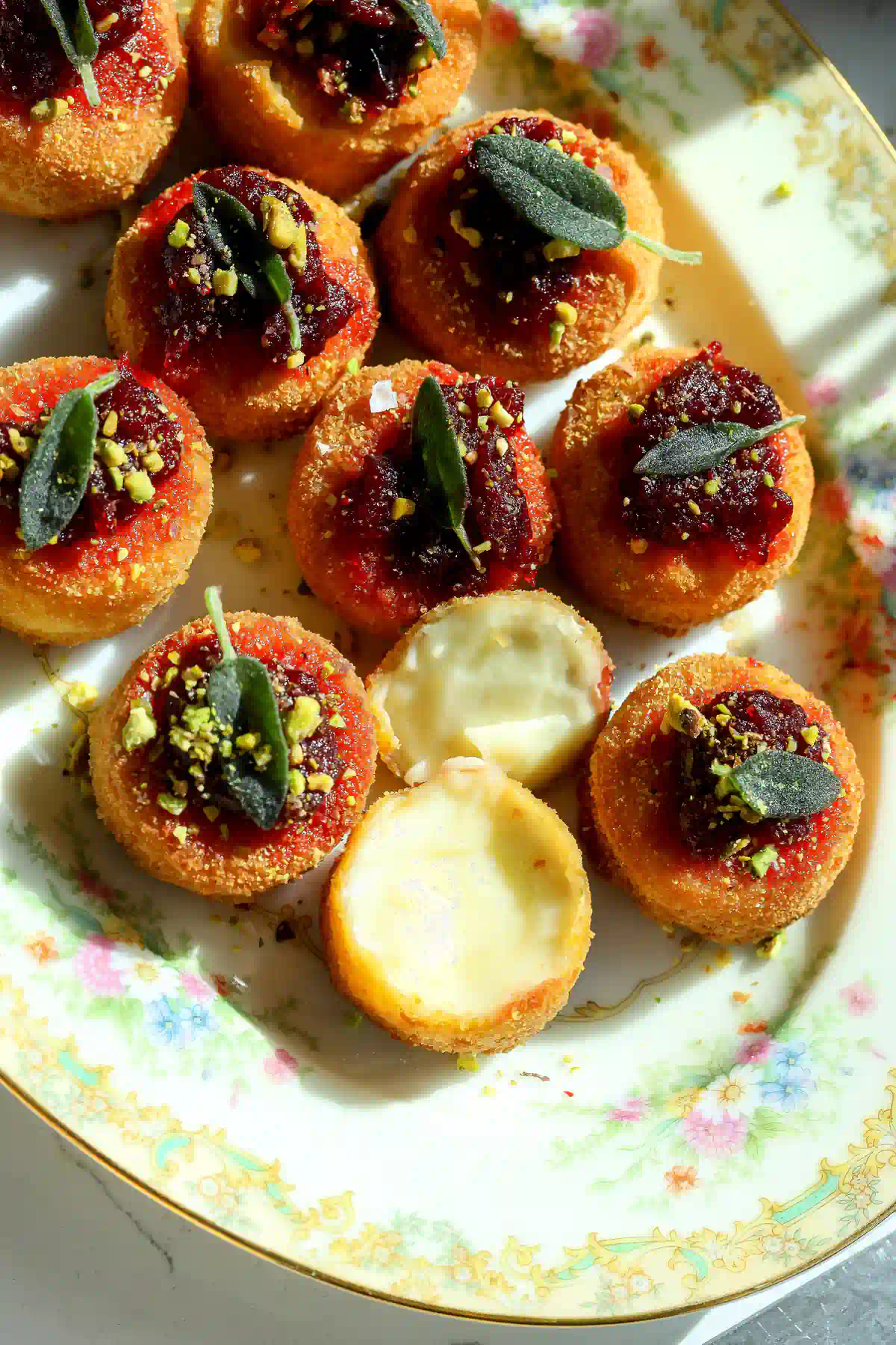Crispy fried brie bites topped with cranberry chipotle sauce, crushed pistachio, and fried sage, two cut open to show the warm melted brie inside.