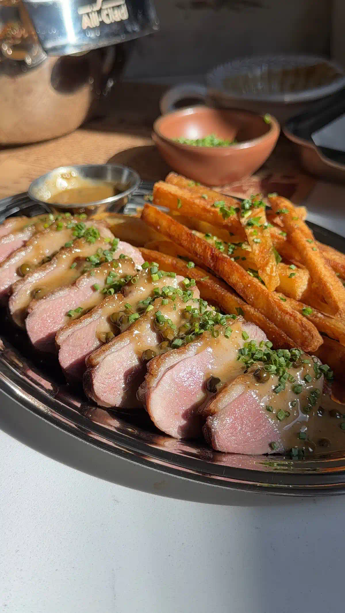 Close-up of medium-rare duck breast slices with crispy skin and rosy pink centers, drizzled with miso ginger pan sauce and chives, frites alongside