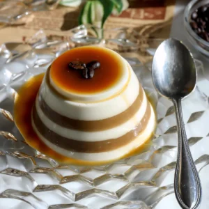An espresso eggnog panna cotta with cream and coffee stripes, caramel dripping down the sides and three coffee beans on top, on a glass plate with a spoon.
