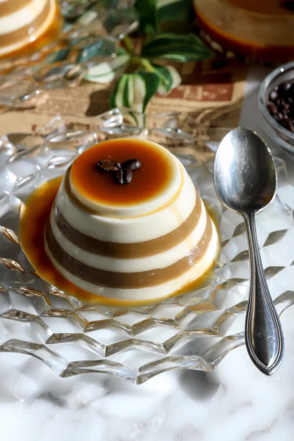 An espresso eggnog panna cotta with cream and coffee stripes, caramel dripping down the sides and three coffee beans on top, on a glass plate with a spoon.