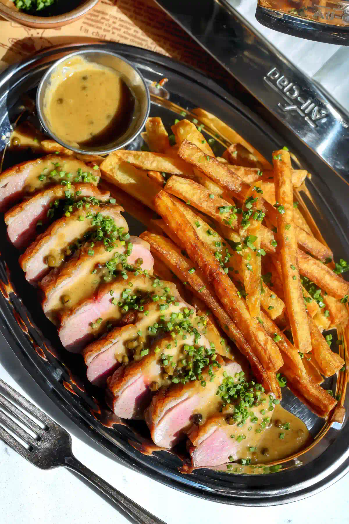 Overhead view of sliced duck breast in miso ginger pan sauce with green peppercorns and chives, next to a pile of golden frites topped with chives