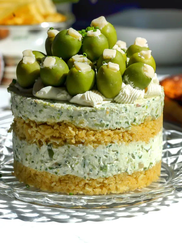 Layered savory dirty martini cheesecake on a glass plate, topped with castelvetrano olives, blue cheese, and chives.