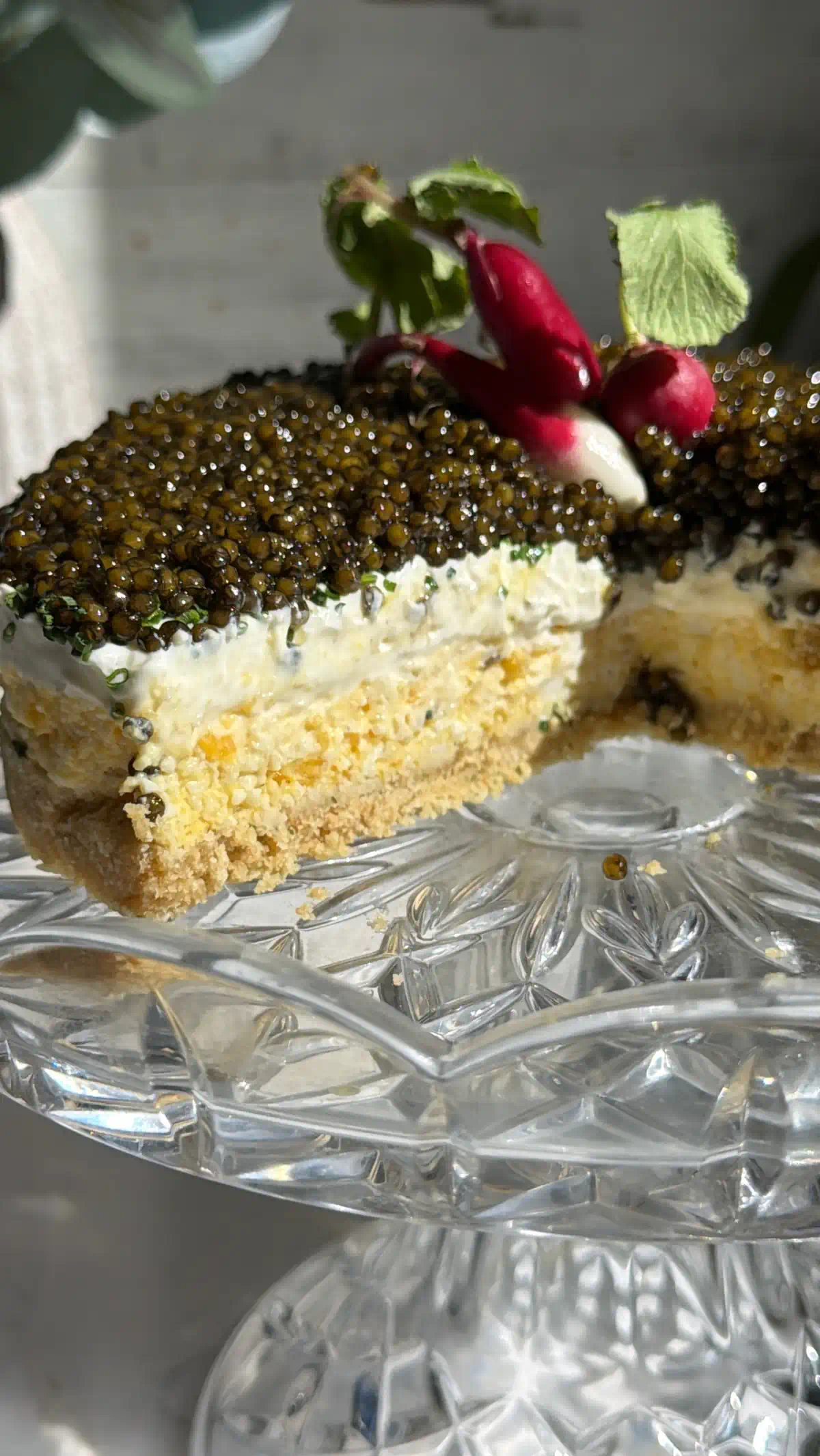Caviar pie on a crystal cake stand with one slice removed, the cut face showing four layers and radishes on top.
