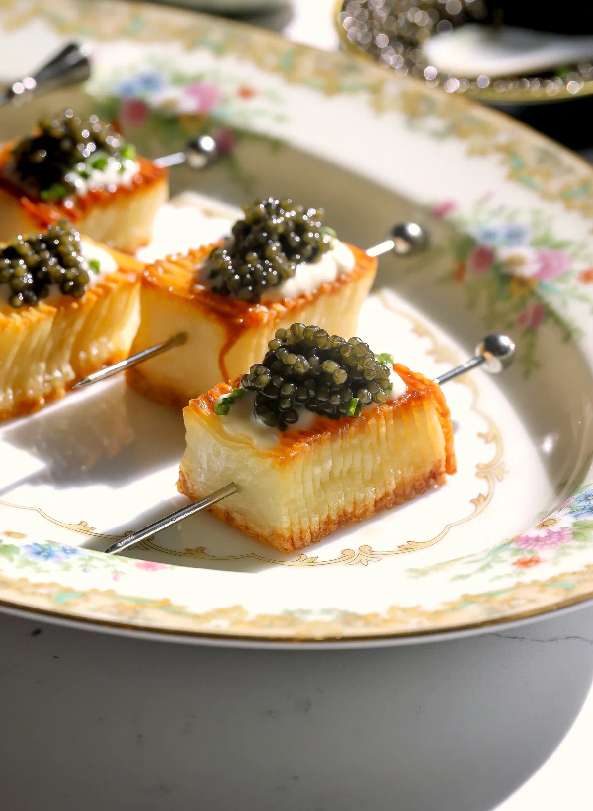 Sunlit potato skewers with golden edges and soft layered centers, topped with caviar, on a floral plate with a tin of caviar behind.